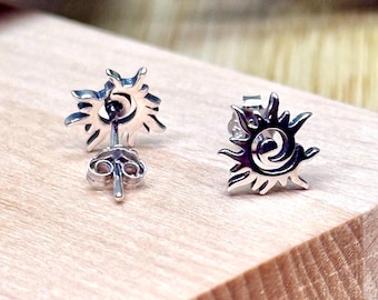 May include: A pair of silver-colored stud earrings. Each earring features a sun design with radiating rays and a central spiral. The earrings are displayed on a light-colored wooden surface.