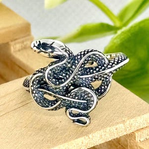 May include: A silver-coloured ring depicting a coiled snake. The snake's head is raised, and its body is textured with scales. The ring is set against a light wooden surface.