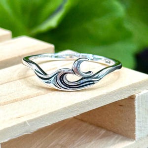 May include: A silver ring with a wave design. The ring features a stylized wave with curved lines and a crescent shape. The ring is set against a wooden surface with a green background.