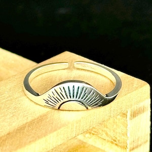 May include: A silver-colored ring with an engraved sunburst design. The ring is adjustable and sits on a wooden surface. The sunburst design features a half-circle with radiating lines, creating a minimalist and stylish accessory.