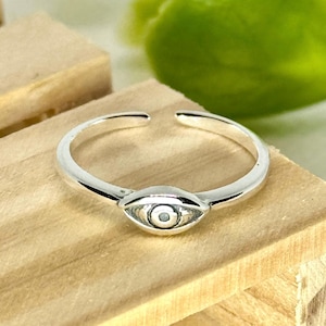 Peut inclure: Bague argentée ornée d'un motif en forme d'œil au centre. La bague est ouverte en haut et repose sur une surface en bois brun clair. Le motif de l'œil présente une pupille et un iris détaillés.