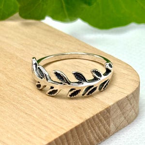 May include: A silver ring featuring a leaf design. The ring has a band and a detailed leaf pattern that wraps around the top. The ring is displayed on a wooden surface, showcasing its intricate design and craftsmanship.