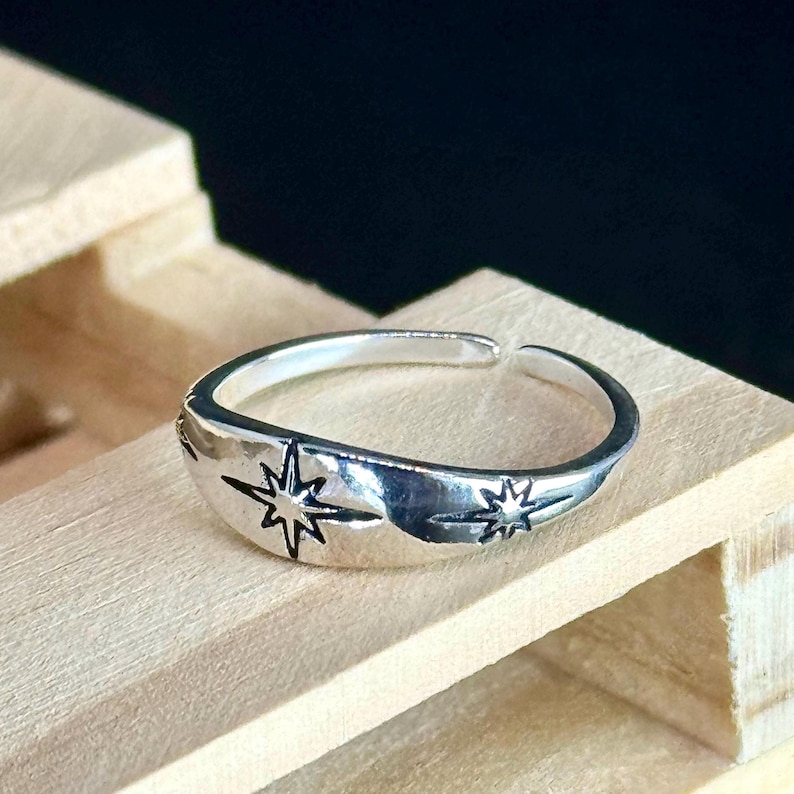 May include: A silver-colored ring with an open design. The ring features two starburst designs. The ring is displayed on a wooden surface.
