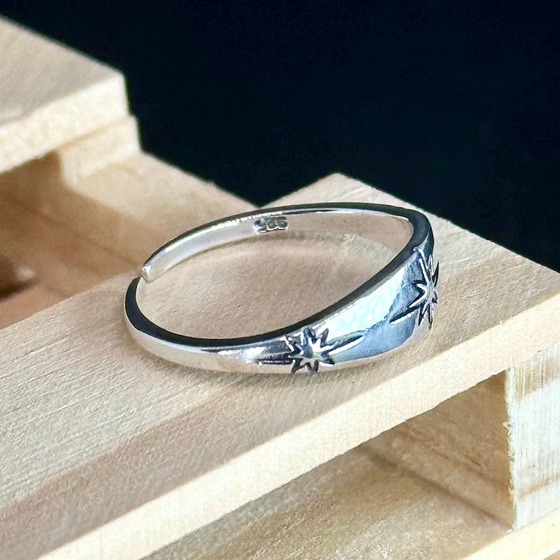May include: A silver-colored ring with a starburst design. The ring is adjustable and features a small star on the band and a larger star on the face. The ring is made of a shiny metal and is displayed on a wooden surface.