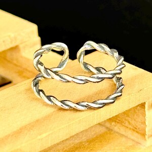 May include: A silver-colored ring with a twisted rope design. The ring has two connected bands, with a textured, braided appearance. The ring is displayed on a light-colored wooden surface, with a dark background.