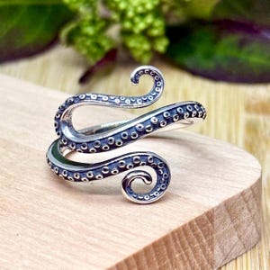 May include: A silver-toned ring in the shape of an octopus tentacle, with detailed suckers. The ring wraps around the finger in a spiral design. The ring is set on a light-colored wooden surface.
