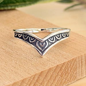 May include: A V-shaped silver ring featuring a pointed chevron design. The band has a dark, oxidized recessed background with a repeating pattern of raised, polished silver U-shaped motifs along the entire outer surface of the ring.