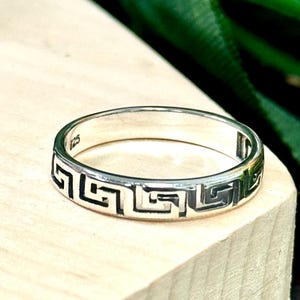 May include: A silver band ring with a Greek key design. The ring is simple and elegant.