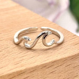 May include: A silver-colored ring with a wave design. The ring is open at the back, allowing for size adjustment. The wave design is a continuous, flowing shape, creating a minimalist and elegant aesthetic. The ring is displayed on a wooden surface.