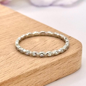May include: A silver ring with a continuous pattern of small, clear gemstones. The ring is set on a light brown wooden surface. The gemstones are oval-shaped and evenly spaced around the band.