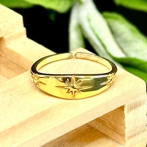 May include: A gold-colored ring with a starburst design. The ring has an open design and is set on a wooden surface. The starburst is engraved on the front of the ring, with additional starburst designs on the sides.