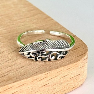 May include: A silver-colored adjustable ring featuring a detailed feather design. The feather has etched lines and decorative scrollwork. The ring is displayed on a wooden surface.