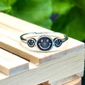 May include: A silver ring featuring three smiley face designs. The central smiley face is larger, with a starburst pattern in the center. The ring is set against a wooden surface with a green background.