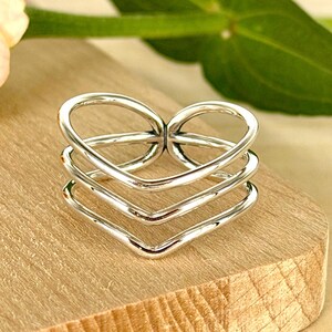 May include: A polished silver-tone ring featuring a multi-layered design. The band consists of three distinct, interconnected wire-like loops that converge to form a heart-shaped silhouette at the top, creating a stacked appearance.