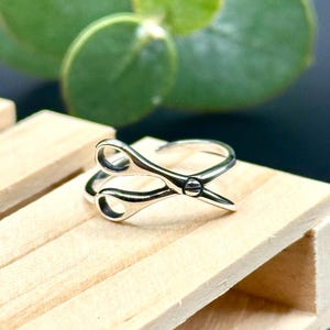 May include: A silver-colored ring shaped like a pair of scissors. The ring is set against a light wooden surface with green foliage in the background. The scissors are detailed with a pivot point and curved handles.