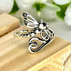 May include: A silver-colored dragonfly ring. The ring features a detailed dragonfly design with openwork wings and a beaded body. The ring is adjustable and sits on a wooden surface.