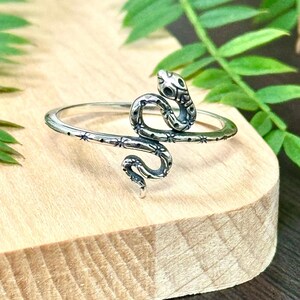 May include: A silver snake ring with a detailed design. The snake is coiled and has a textured surface. The ring is on a wooden surface.