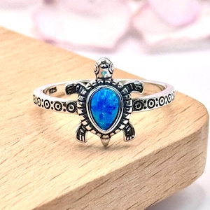 May include: A silver ring featuring a turtle design with a vibrant blue opal stone. The turtle's shell and body are detailed with intricate patterns. The ring band has a repeating pattern of circles and dots.