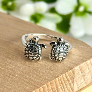 May include: A silver-toned ring featuring two detailed turtle figures facing each other. The turtles have textured shells and small flipper details. The ring is set on a wooden surface, with blurred green and white foliage in the background.