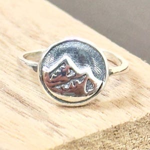 May include: A silver ring featuring a circular design with a mountain range. The ring has a detailed mountain scene in the center, with a smooth, polished band. The ring is set on a wooden surface.