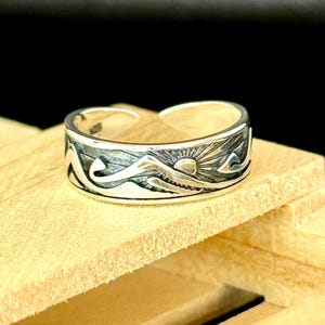 May include: A silver ring with a mountain and sun design. The ring features a detailed landscape with mountains, waves, and a sunburst. The ring is set on a wooden surface.