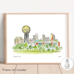 Puede incluir: Una ilustración de acuarela del horizonte de Knoxville, TN, con el Sunsphere, un gran edificio con forma de esfera. La ilustración está en un marco blanco con un borde de madera claro.