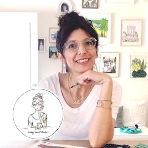 Op de afbeelding: Een vrouw met donker haar dat in een knotje is geknoopt, draagt een bril en een wit shirt. Ze zit aan een tafel met een wit oppervlak. Ze houdt een pen in haar rechterhand en haar linkerhand rust op haar kin. Ze glimlacht naar de camera. Er zit een ronde sticker op de tafel met een zwart-witte illustratie van een vrouw met een bril en een knotje. De sticker zegt "messy heart studio".