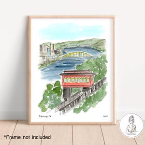 Peut inclure: Impression aquarelle d'un paysage urbain de Pittsburgh, PA. L'œuvre représente un funiculaire rouge, un pont jaune et des bâtiments au bord d'une rivière. Le ciel est bleu clair et le paysage environnant est vert.