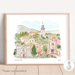 May include: Watercolor print of Santa Fe, NM, featuring a detailed depiction of buildings, a church, and a tower, set against a backdrop of green hills and a light blue sky. The artwork is framed in a light wood frame.