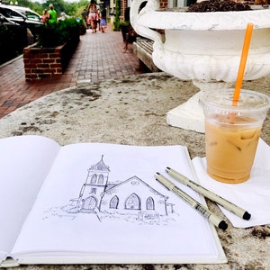 May include: A black and white line drawing of a church building on a white sketchbook. The sketchbook is open and lying on a table with a glass of iced coffee and two black pens.