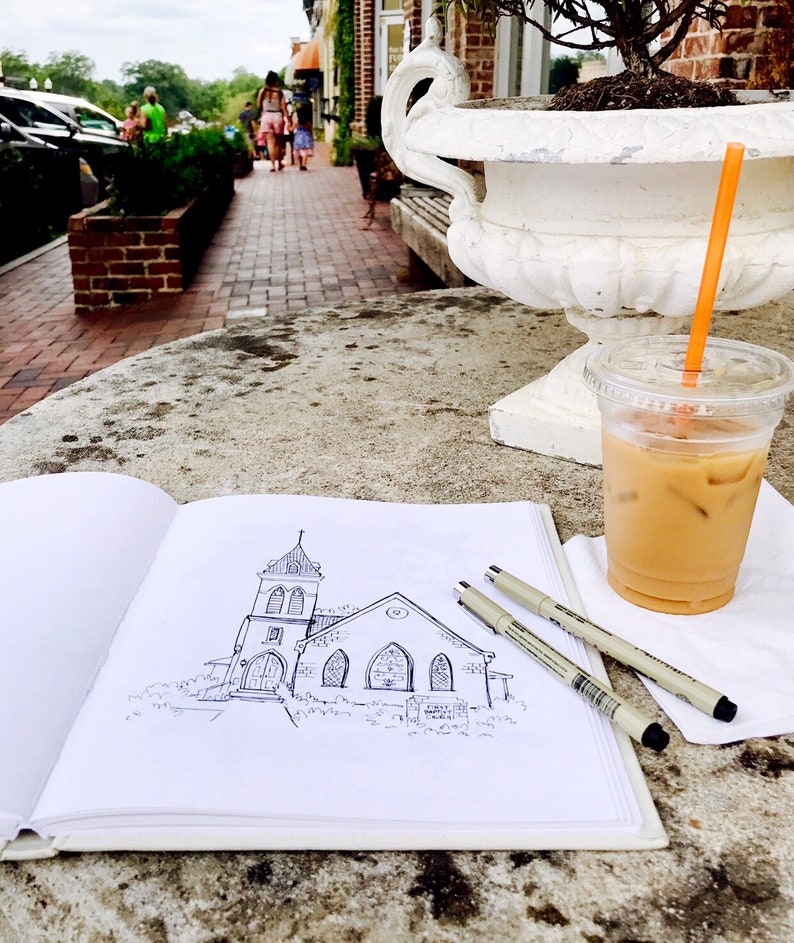 May include: A black and white line drawing of a church building on a white sketchbook. The sketchbook is open and lying on a table with a glass of iced coffee and two black pens.