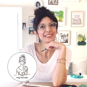 May include: A woman with dark hair pulled back in a bun is wearing glasses and a white shirt. She is sitting at a table with a white surface. She is holding a pen in her right hand and her left hand is resting on her chin. She is smiling at the camera. There is a round sticker on the table with a black and white illustration of a woman with glasses and a bun. The sticker says "messy heart studio".