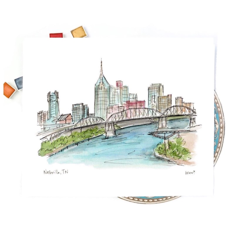 K&ouml;nnte beinhalten: Ein Aquarellbild der Skyline von Nashville mit einer Br&uuml;cke &uuml;ber einen Fluss. Das Bild ist in Blaut&ouml;nen, Gr&uuml;n- und Braunt&ouml;nen gehalten. Der Text "Nashville, TN" steht in der linken unteren Ecke.