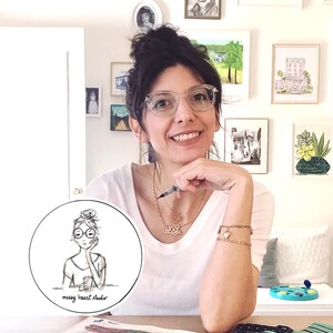 Puede incluir: Una mujer con el pelo oscuro recogido en un mo&ntilde;o lleva gafas y una camisa blanca. Est&aacute; sentada en una mesa con una superficie blanca. Sostiene un bol&iacute;grafo en su mano derecha y su mano izquierda descansa sobre su barbilla. Sonr&iacute;e a la c&aacute;mara. Hay una pegatina redonda en la mesa con una ilustraci&oacute;n en blanco y negro de una mujer con gafas y un mo&ntilde;o. La pegatina dice "messy heart studio".