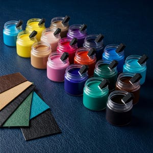 May include: A collection of small glass jars filled with various colors of paint, each with a black sponge applicator. Leather swatches in shades of brown, green, and blue are arranged on a dark blue surface.