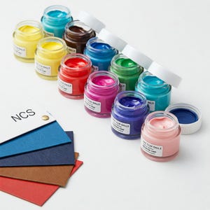 Peut inclure: Un ensemble de 10 petits pots en verre avec couvercles, remplis de peinture de différentes couleurs. Les pots sont disposés en rangée sur une surface blanche. La peinture est étiquetée "NCS Color Balls Acrylic Lacquer".