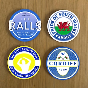 May include: Four round coasters with different designs. The first coaster is blue with white text that reads "Captain Fantastic Joe Ralls Born in Aldershot Made in Cardiff". The second coaster is white with a red dragon and green and white stripes. The text reads "Pride of South Wales Cardiff". The third coaster is yellow with a white silhouette of a person with their arms raised. The text reads "Do the Ayatollah It's a Cardiff Thing". The fourth coaster is blue with a white bird and two crossed flags. The text reads "Cardiff 1899".