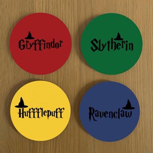 May include: Four round coasters featuring the house names from the Harry Potter series. The coasters are red, green, yellow, and blue. Each coaster has a black silhouette of a witch's hat and the house name in black lettering. The house names are Gryffindor, Slytherin, Hufflepuff, and Ravenclaw.