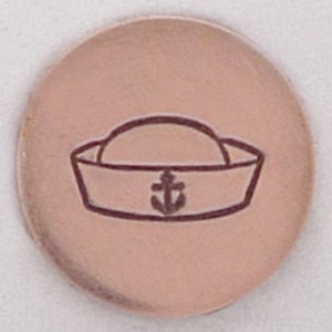 May include: A round, copper-colored metal charm with a sailor hat and anchor design.