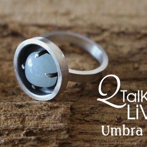 Può includere: Un anello d'argento con una pietra preziosa blu chiaro incastonata in una lunetta circolare. L'anello è su una superficie di legno.  "Q Talk LIVE" e "Umbra Ring" sono scritti in bianco sulla superficie.