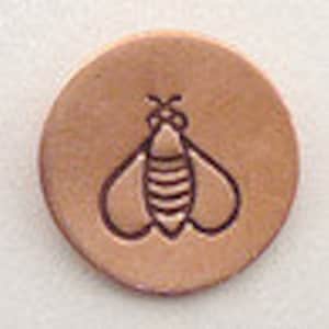 May include: A copper-colored metal disc with a simple line drawing of a bee in the center.