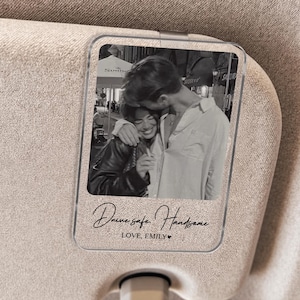 May include: Clear acrylic car visor clip featuring a black and white photo of a couple embracing. The clip has the text "Drive safe, Handsome. LOVE, EMILY" in a handwritten style font. The clip is attached to a beige car visor.