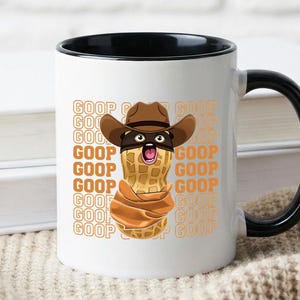 May include: White ceramic mug with a black handle and interior. The mug features a cartoon peanut character wearing a cowboy hat and bandana, with the word "GOOP" repeated in an orange font.