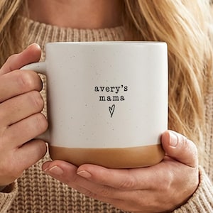 Op de afbeelding: Een gespikkelde witte keramische mok met een bruine basis, met de tekst "avery's mama" en een hartsymbool. De mok wordt vastgehouden door een persoon die een beige trui draagt.
