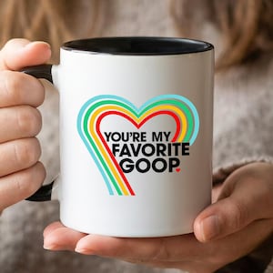 Könnte beinhalten: Weiße Keramiktasse mit schwarzem Innenraum und Henkel. Die Tasse hat ein farbenfrohes Herzdesign mit dem Text "YOU'RE MY FAVORITE GOOP."