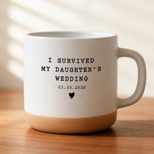 May include: A white ceramic mug with a speckled finish and a tan base. The mug has the text "I SURVIVED MY DAUGHTER'S WEDDING 03.03.2026" with a black heart symbol. The mug has a large handle.