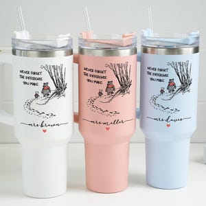Op de afbeelding: Drie geïsoleerde bekers in wit, roze en lichtblauw, elk met een handvat en een rietje. De bekers hebben een zwart-wit illustratie met de tekst "NEVER FORGET THE DIFFERENCE YOU MAKE" en een naam eronder.
