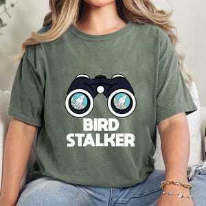 May include: A green t-shirt with a white graphic of binoculars with two birds in the lenses. The text "BIRD STALKER" is below the binoculars.