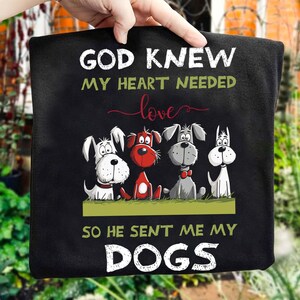 A person is holding a black t-shirt with a graphic design of four cartoon dogs and text that reads "God Knew my heart needed love so he sent me my dogs".