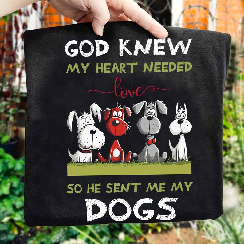 A person is holding a black t-shirt with a graphic design of four cartoon dogs and text that reads "God Knew my heart needed love so he sent me my dogs".
